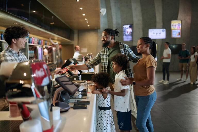 Padres afroamericanos felices y sus hijos comprando entradas en el puesto de comida de un estadio deportivo.